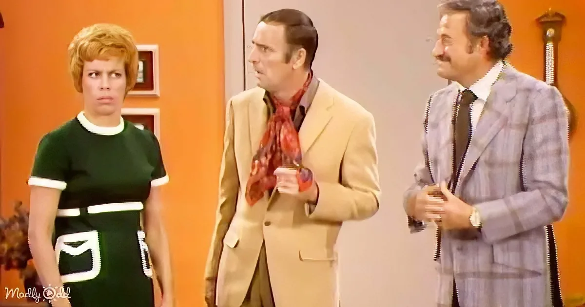 A woman in a green dress stands next to a man in a tan suit and another man in a striped suit, all in a room with orange...