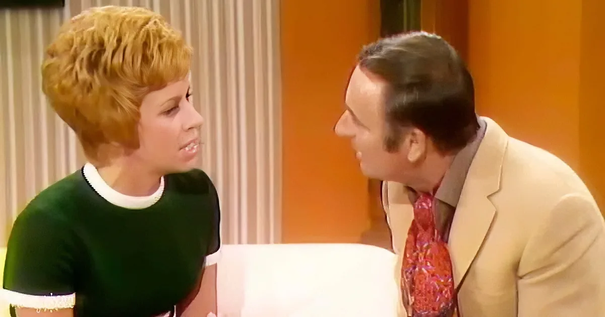 A woman with short blonde hair and a green dress is talking to a man in a beige suit and red tie.