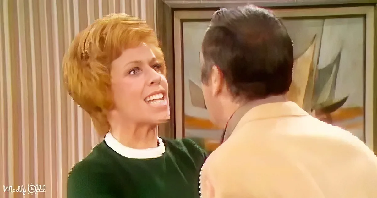 A woman with short red hair, wearing a green sweater, is speaking to a man in a light-colored suit.