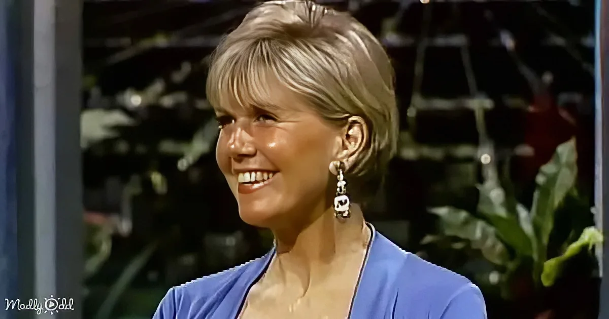 A smiling woman with short blonde hair, wearing a blue top and earrings, stands in front of a blurred background with plants.