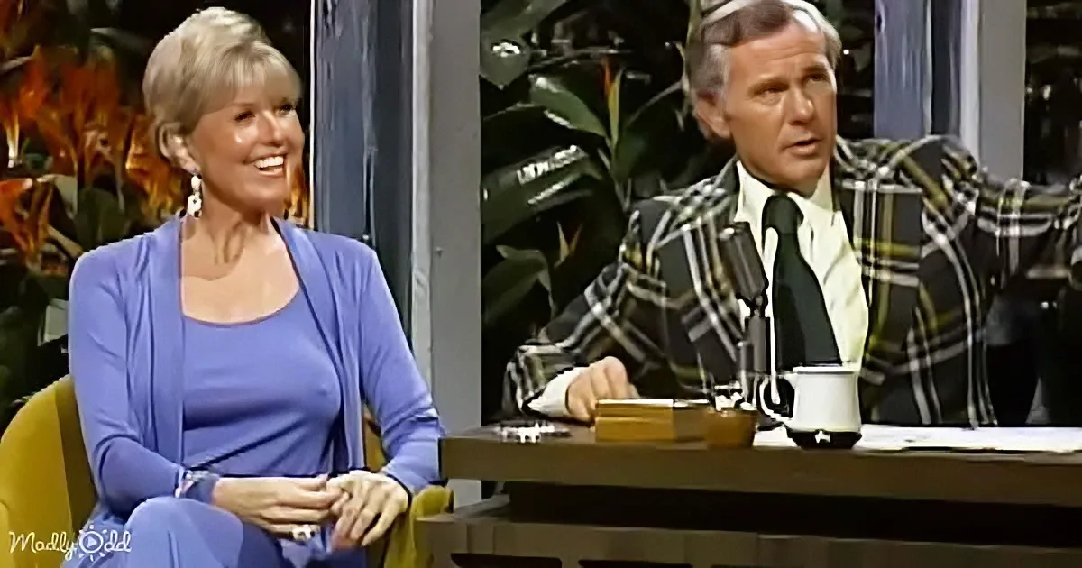 A woman in a blue outfit smiles while seated next to a man in a plaid suit at a desk with a microphone.