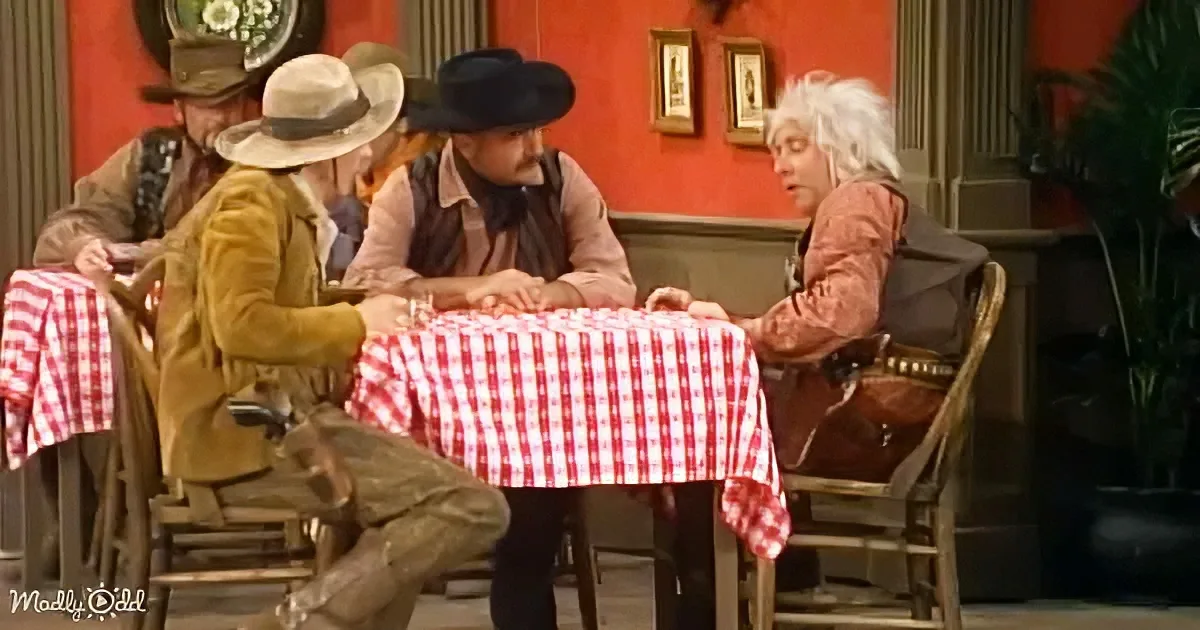 A man in a cowboy hat sits at a table with a red-and-white checkered tablecloth, surrounded by others in period attire.