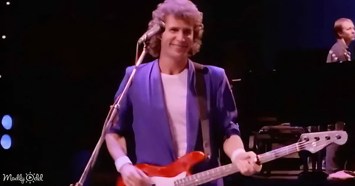 A man in a blue jacket plays a red guitar on stage, smiling near a microphone.