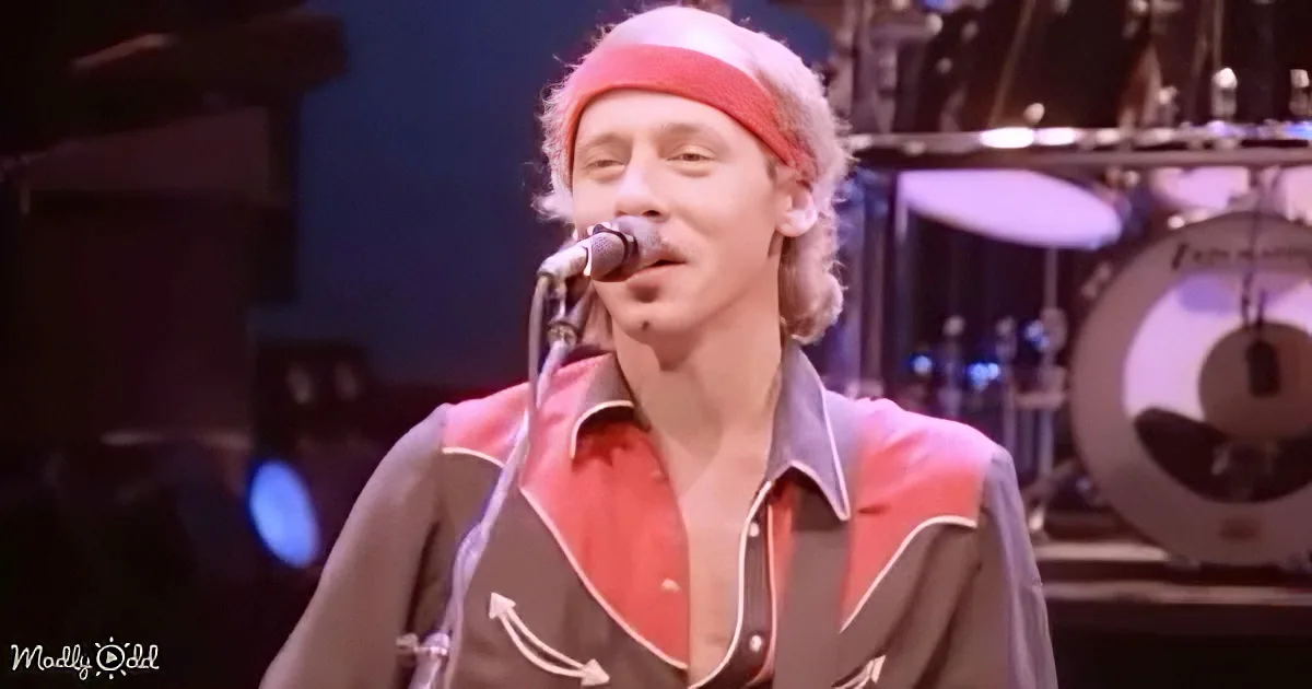 A man in a red and black shirt sings into a microphone on stage with a drum set in the background.
