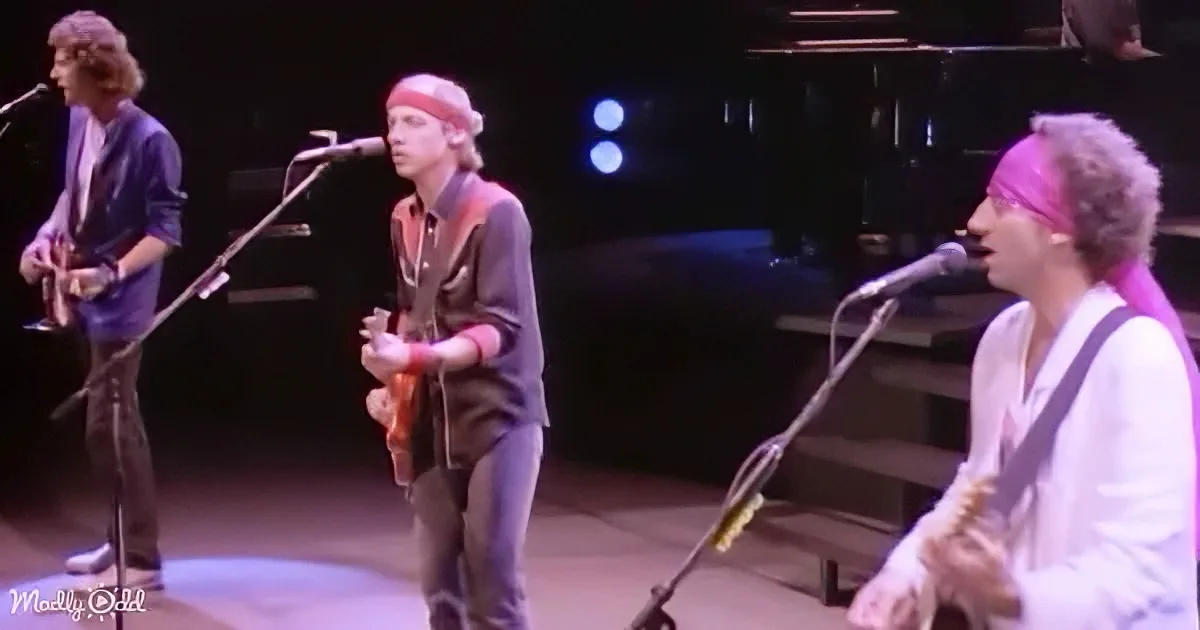 A musician in a red bandana plays guitar on stage with other band members.