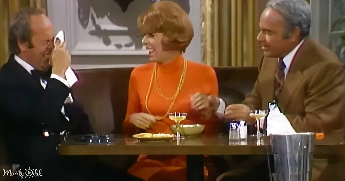 A woman in an orange dress and pearls sits at a table with two men, one wiping his eye, in a warmly lit room.