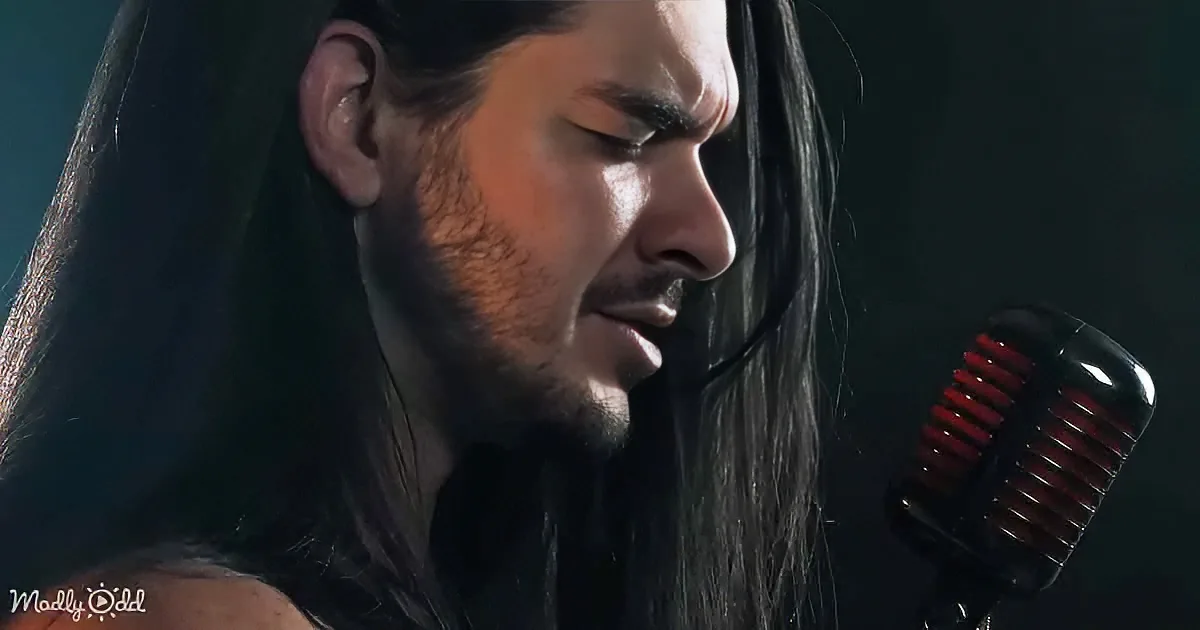 A man with long dark hair and a beard sings into a red microphone, eyes closed, in a dimly lit setting.