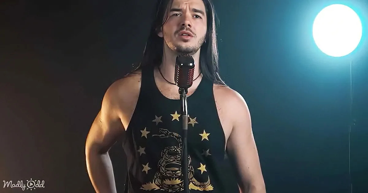 A man with long hair and a black tank top stands on stage, singing into a microphone under bright lighting.