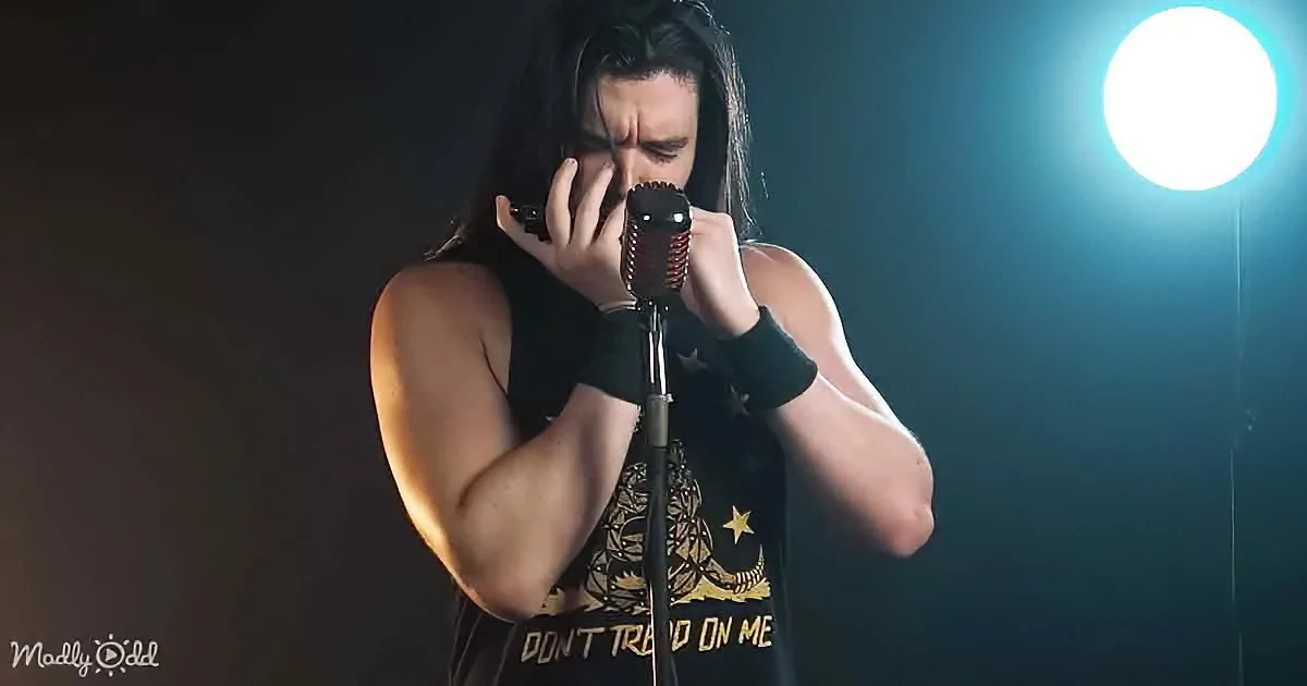 A person in a black tank top with "Don't Tread On Me" sings passionately into a microphone under a spotlight.