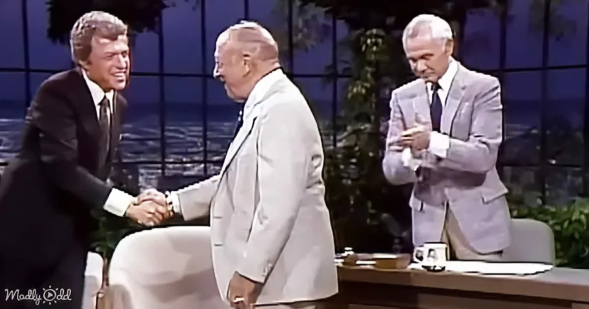 A man in a suit shakes hands with another, while a third man in a suit watches, on a stage with a desk and mug.