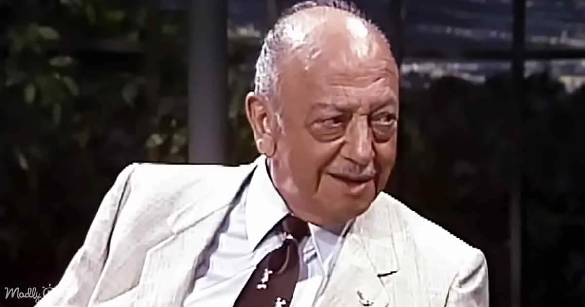An older man in a white suit jacket and tie sits, looking slightly to the side with a calm expression.