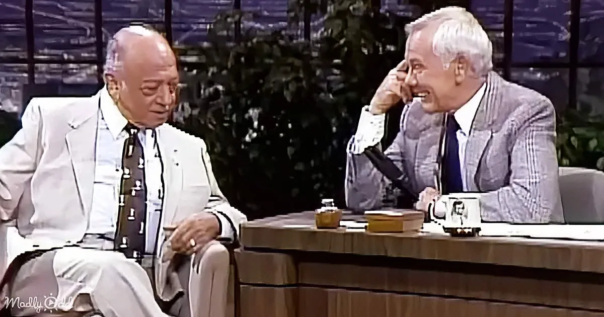 Two men in suits are seated, one laughing, on a talk show set with a desk and mug.