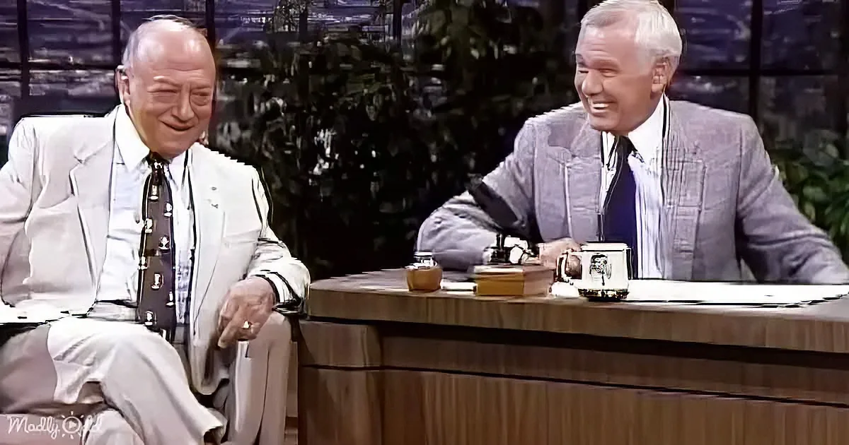 Two men in suits sit at a desk, smiling and laughing, with a mug and books on the desk.
