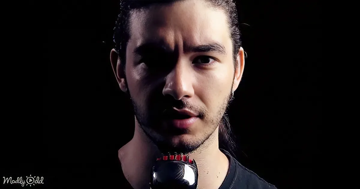 A man with long hair and a beard holds a black object with red accents, set against a dark background.