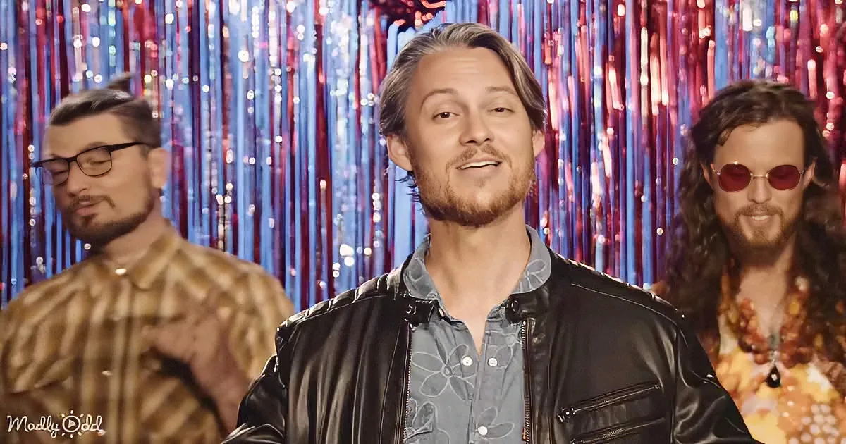 A man in a black leather jacket smiles in front of a colorful, shiny backdrop with two other individuals behind him.