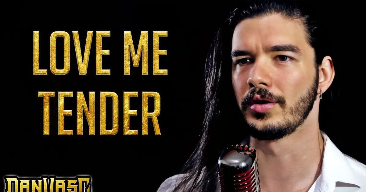A man with long dark hair, a beard, and mustache is holding a microphone, wearing a white shirt, against a dark background.