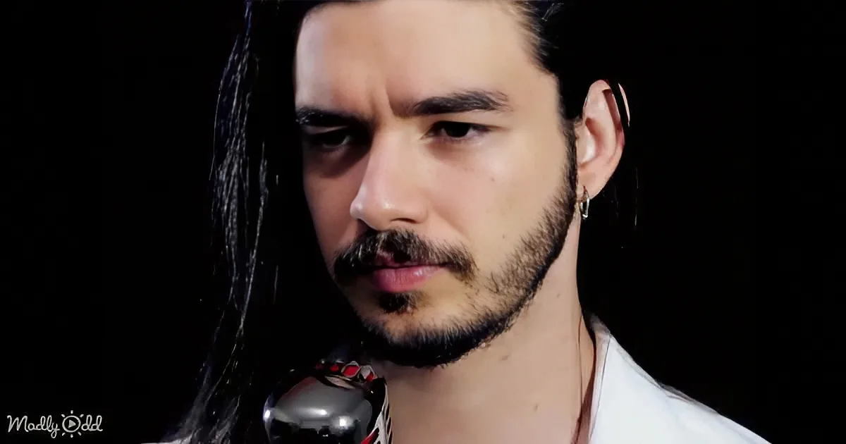 A man with long dark hair, a beard, and a nose ring holds a microphone against a dark background.