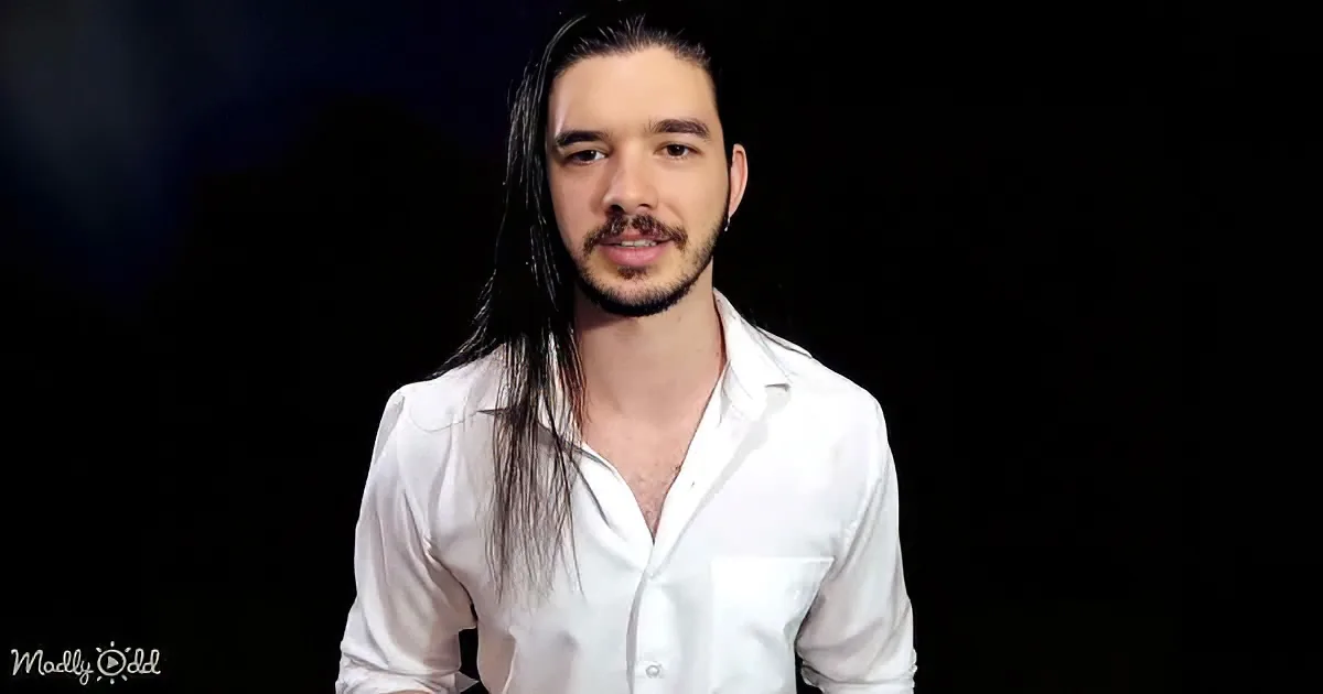 A man with long hair and a beard, wearing a white shirt, stands against a dark background.