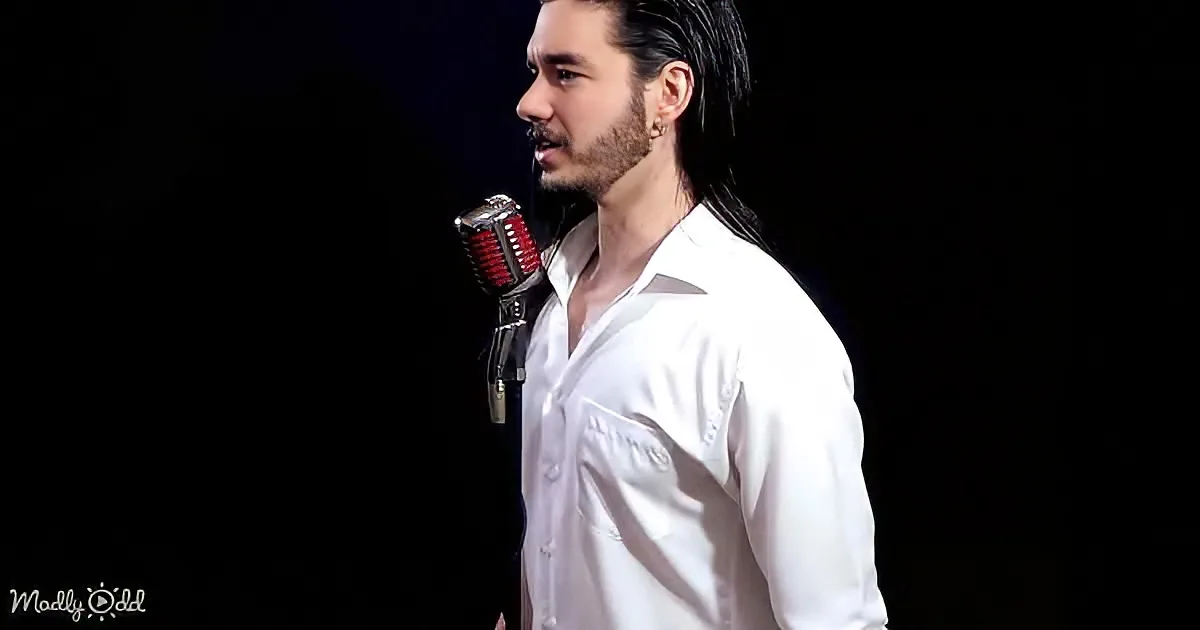 A man in a white shirt sings into a vintage microphone against a dark background.
