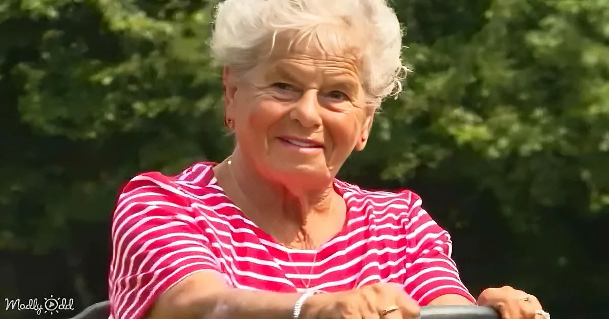 An elderly woman with white hair, wearing a red and white striped shirt, smiles while sitting outdoors with trees in the...