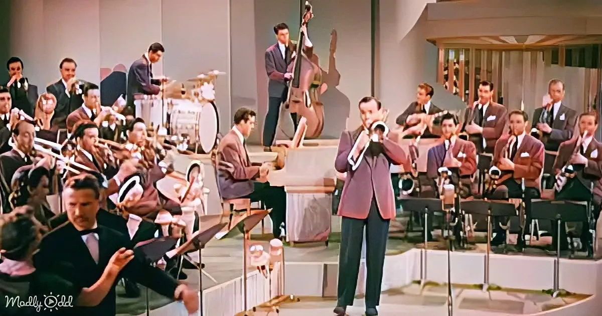 A man in a suit plays a trumpet in front of a large orchestra.