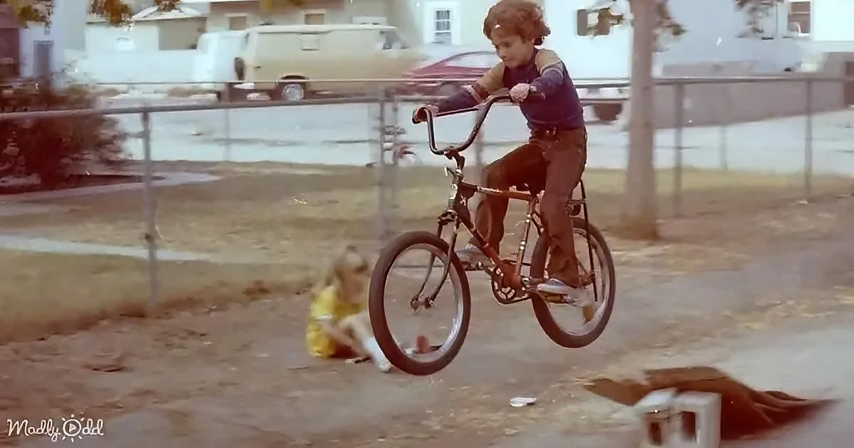 A child rides a bicycle mid-air, performing a wheelie, with another child watching on the ground.