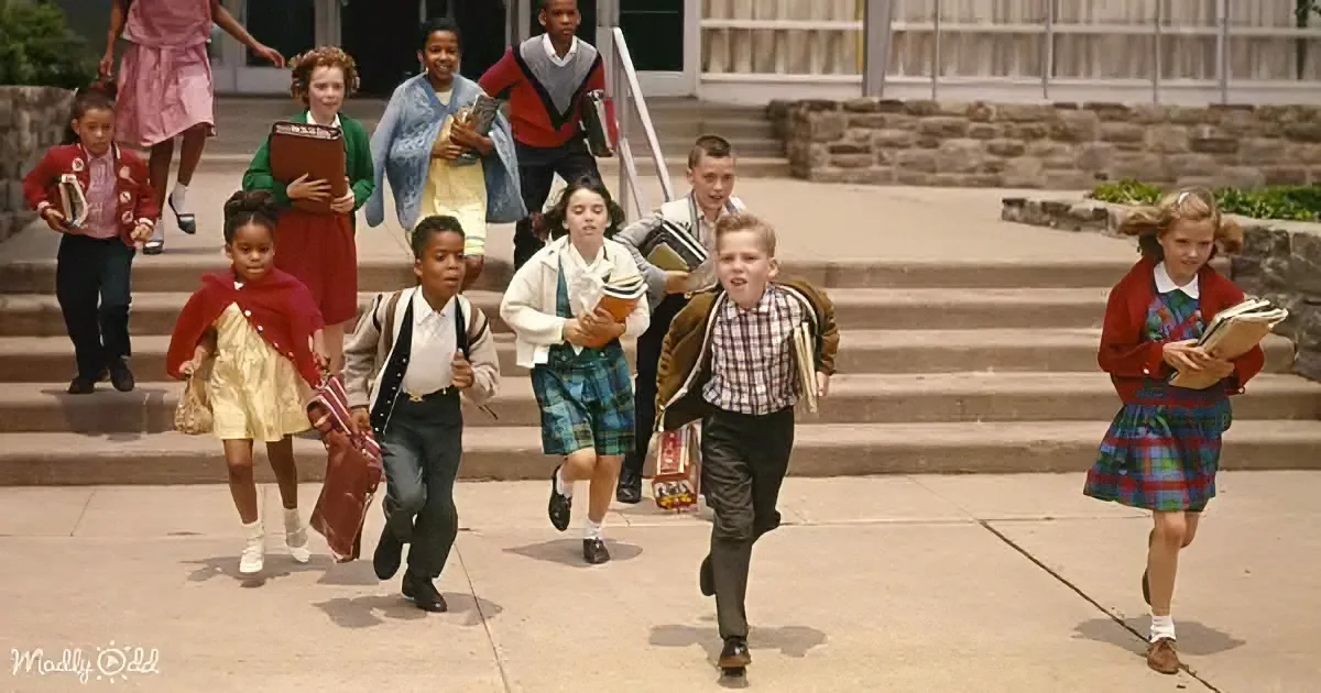 Children in school uniforms run excitedly down steps, holding books and bags, outside a school building.