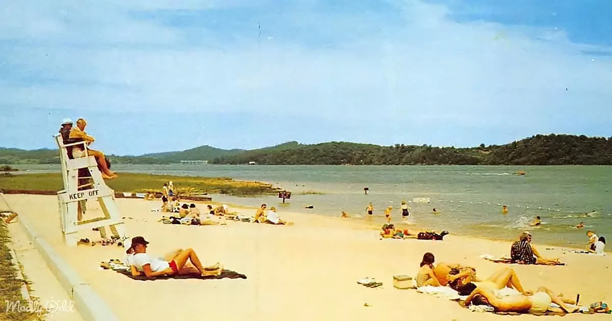 A lifeguard stands on a tower while people relax and swim on a sunny beach.