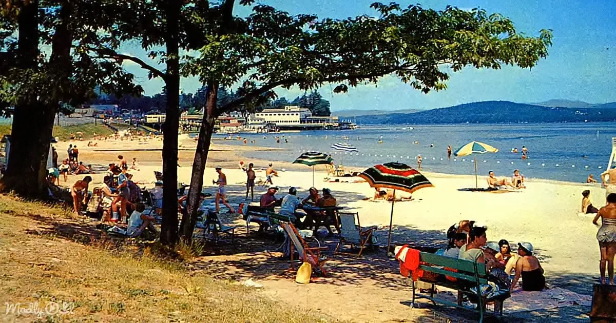 A vibrant beach scene with people relaxing under umbrellas and trees, with a distant ferry and hills in the background.