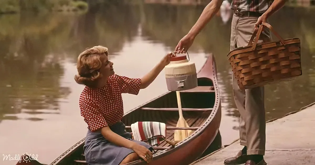 A woman in a red polka-dot shirt and blue skirt sits in a canoe, reaching for a cooler while a man stands nearby holding...