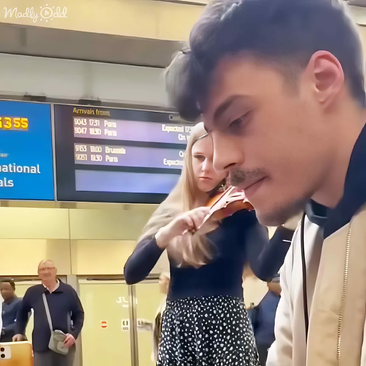 A man in a beige jacket is eating a sandwich at an airport with a woman in a polka dot skirt standing nearby.