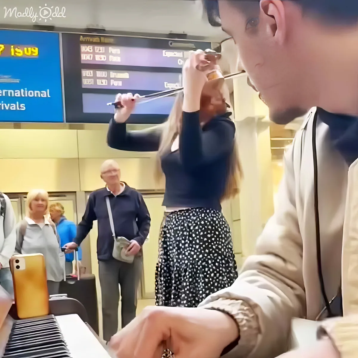 A woman in a polka-dot skirt plays a violin at an airport, surrounded by travelers.