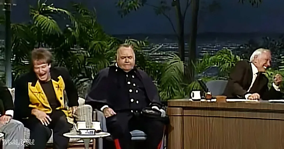 A man in a black military-style jacket sits on a chair, flanked by others in a studio setting with palm trees in the...