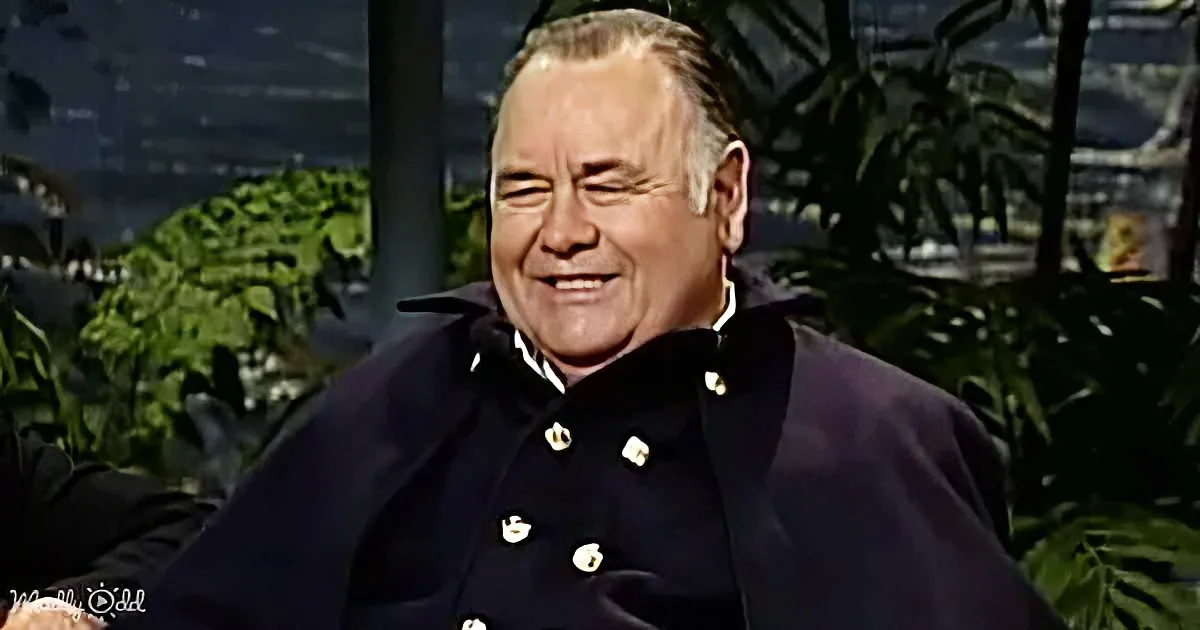 A man in a dark uniform with white buttons smiles, surrounded by greenery and a blurred background.