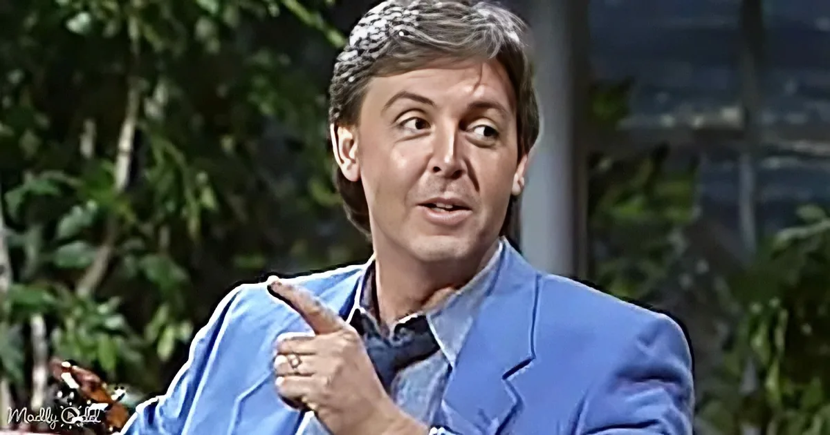 A man in a blue suit points while speaking, surrounded by greenery.