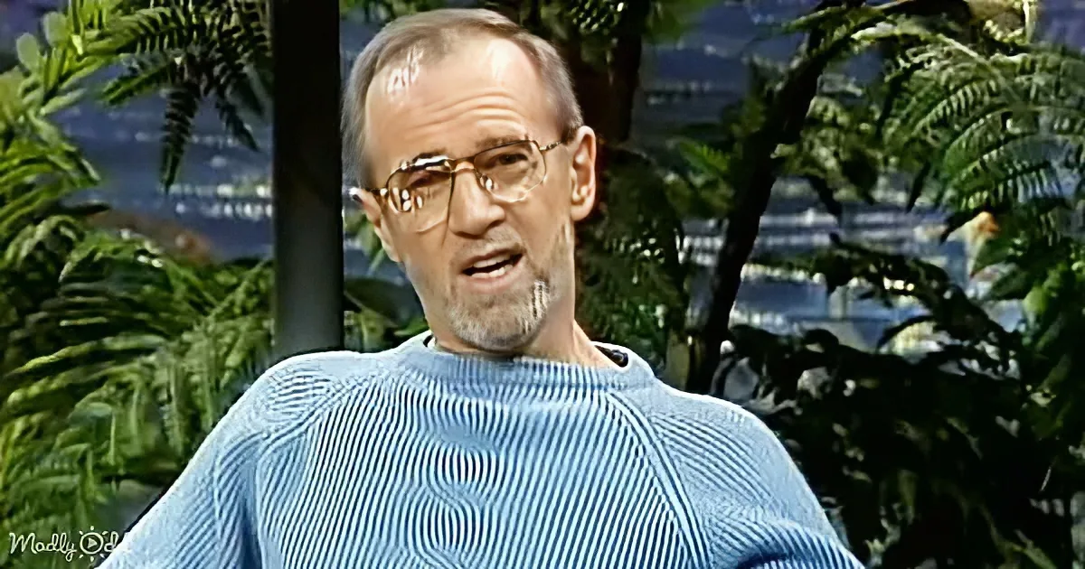 A man in a blue striped sweater speaks, surrounded by green foliage and a blurred background.