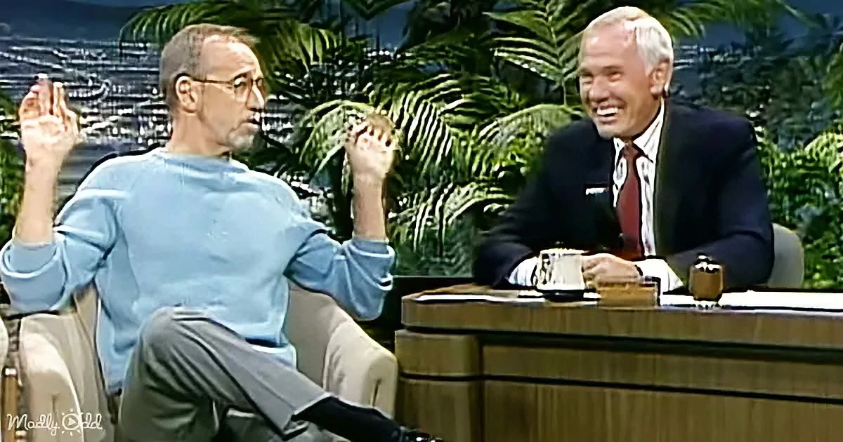A man in a blue sweater gestures animatedly while seated next to a man in a suit on a talk show set.