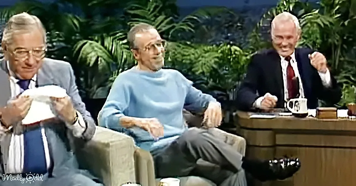 Three men are seated, one in a blue sweater, another in a suit, and the third in a suit, with a tropical backdrop.