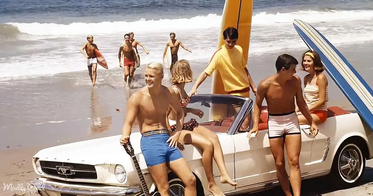A group of people in swimwear on a beach with surfboards and a vintage white car.