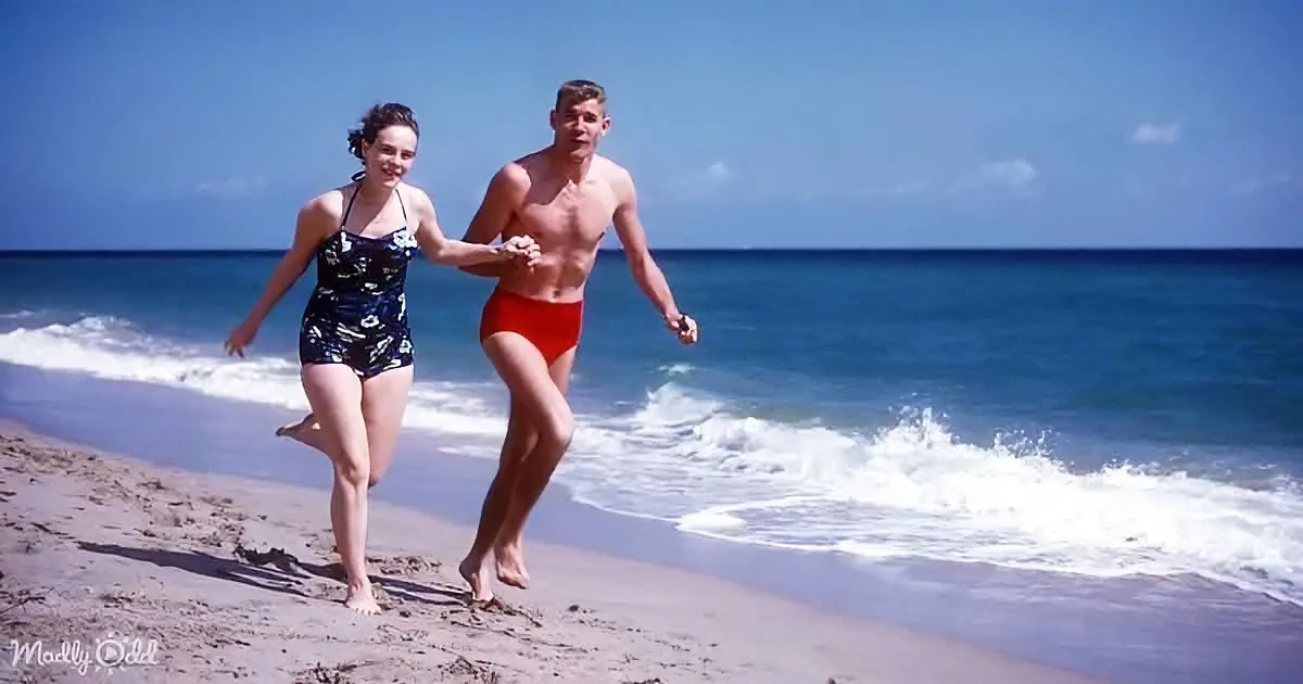 A man and woman in swimsuits run hand-in-hand along a sandy beach towards the ocean.