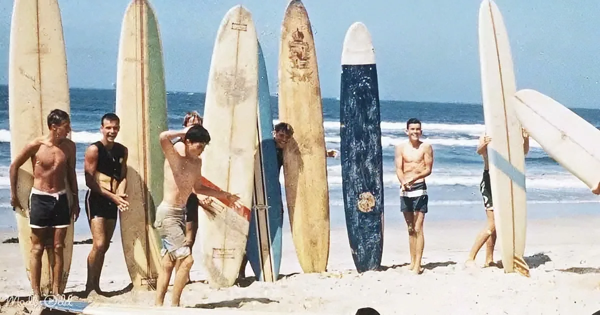 Six surfers with boards on beach, ocean waves in background, sunny day.