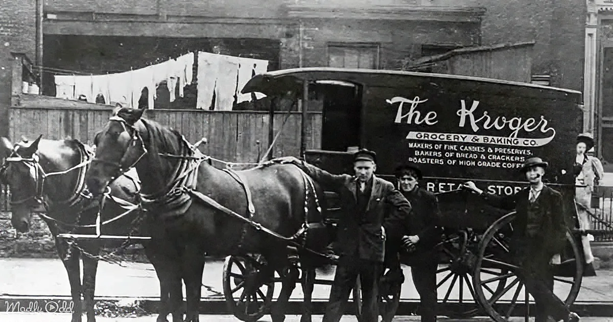 A vintage horse-drawn delivery wagon for The Kroger Grocery & Baking Co. stands in front of a brick building with laundry...
