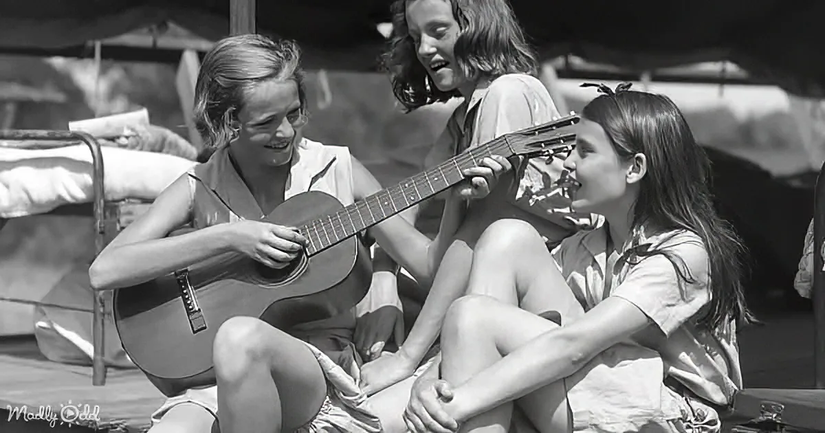 A woman plays guitar while two others watch and smile, sitting outdoors.