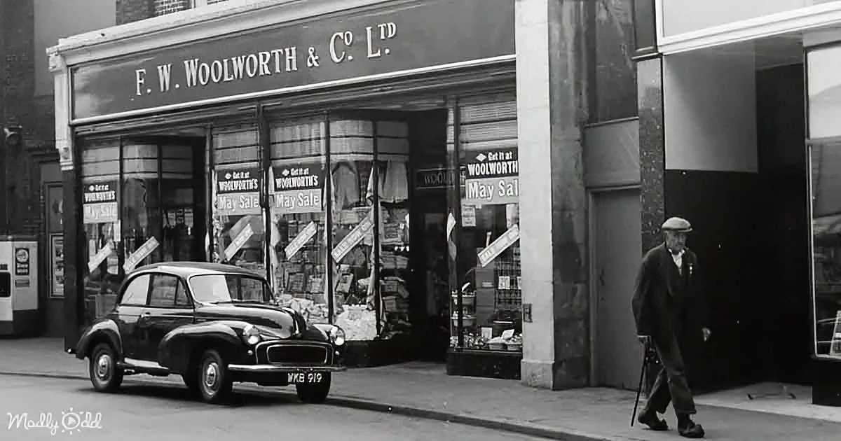 An old man walks past a Woolworths store with a vintage car parked in front.