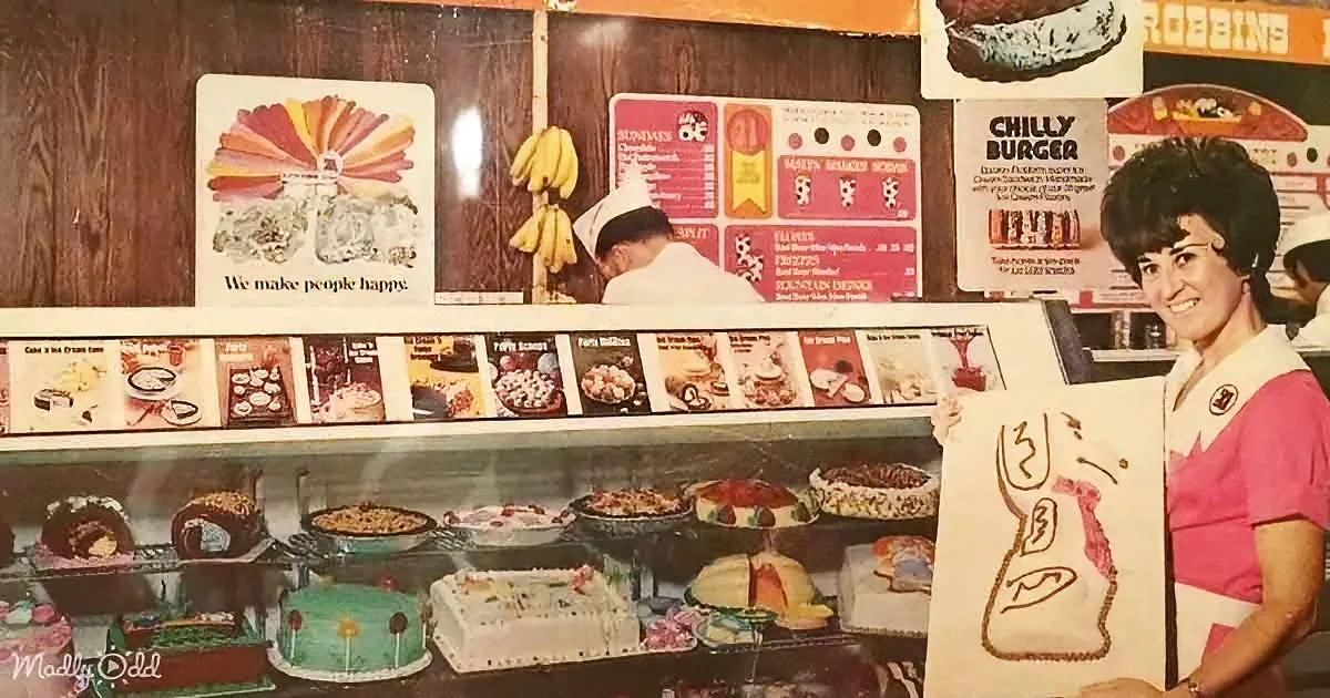 A woman in a pink uniform holds a sign in front of a display case filled with cakes and desserts.