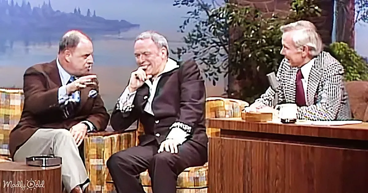 Three men in suits sit on a talk show set, laughing and gesturing animatedly.