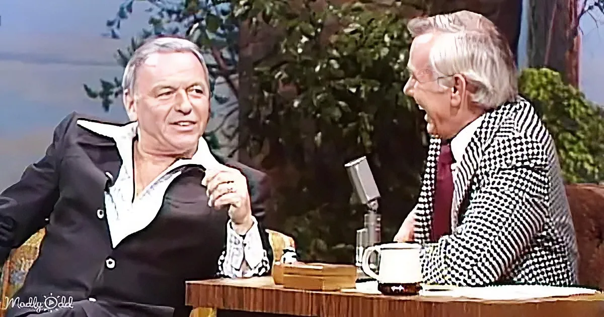 Two men are seated, one in a black suit, the other in a checkered jacket, engaged in conversation on a talk show set.