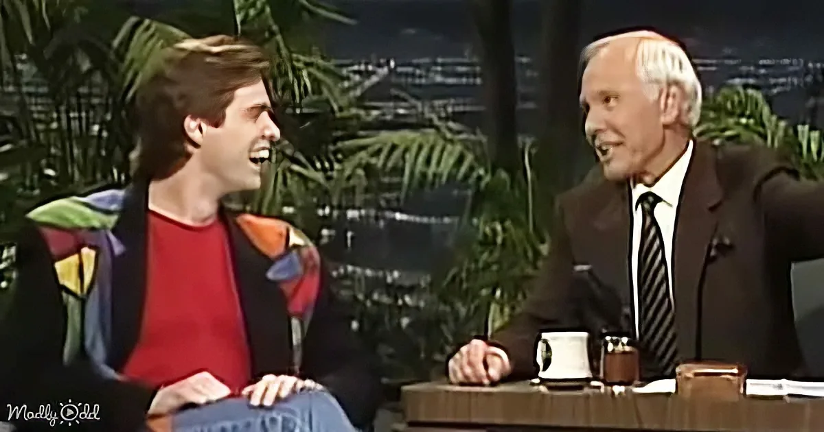 A man in a red shirt and jeans laughs with a suited man in a studio setting.