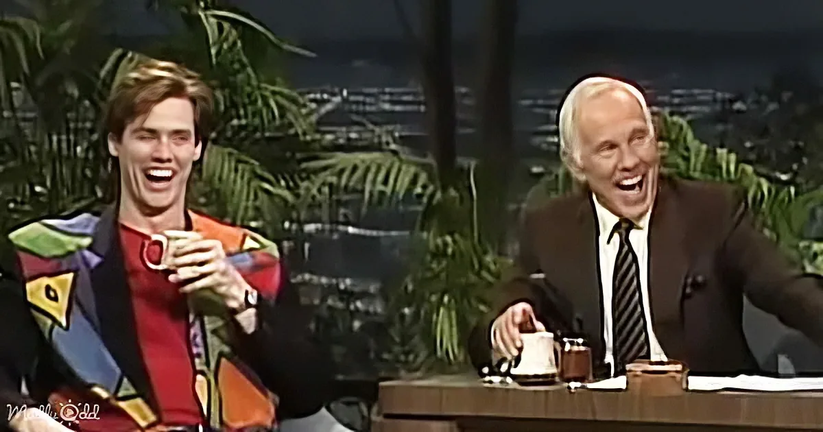 Two individuals are laughing on a talk show set with a palm tree backdrop.