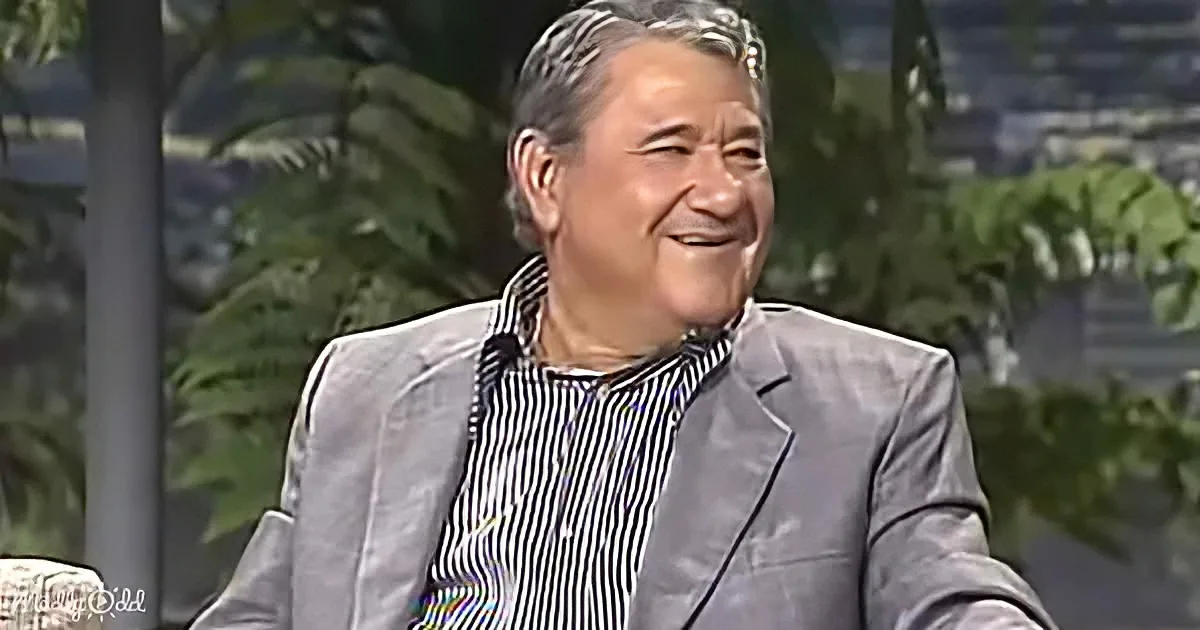 A man in a striped shirt and gray blazer smiles, seated with a blurred background of greenery and a cityscape.